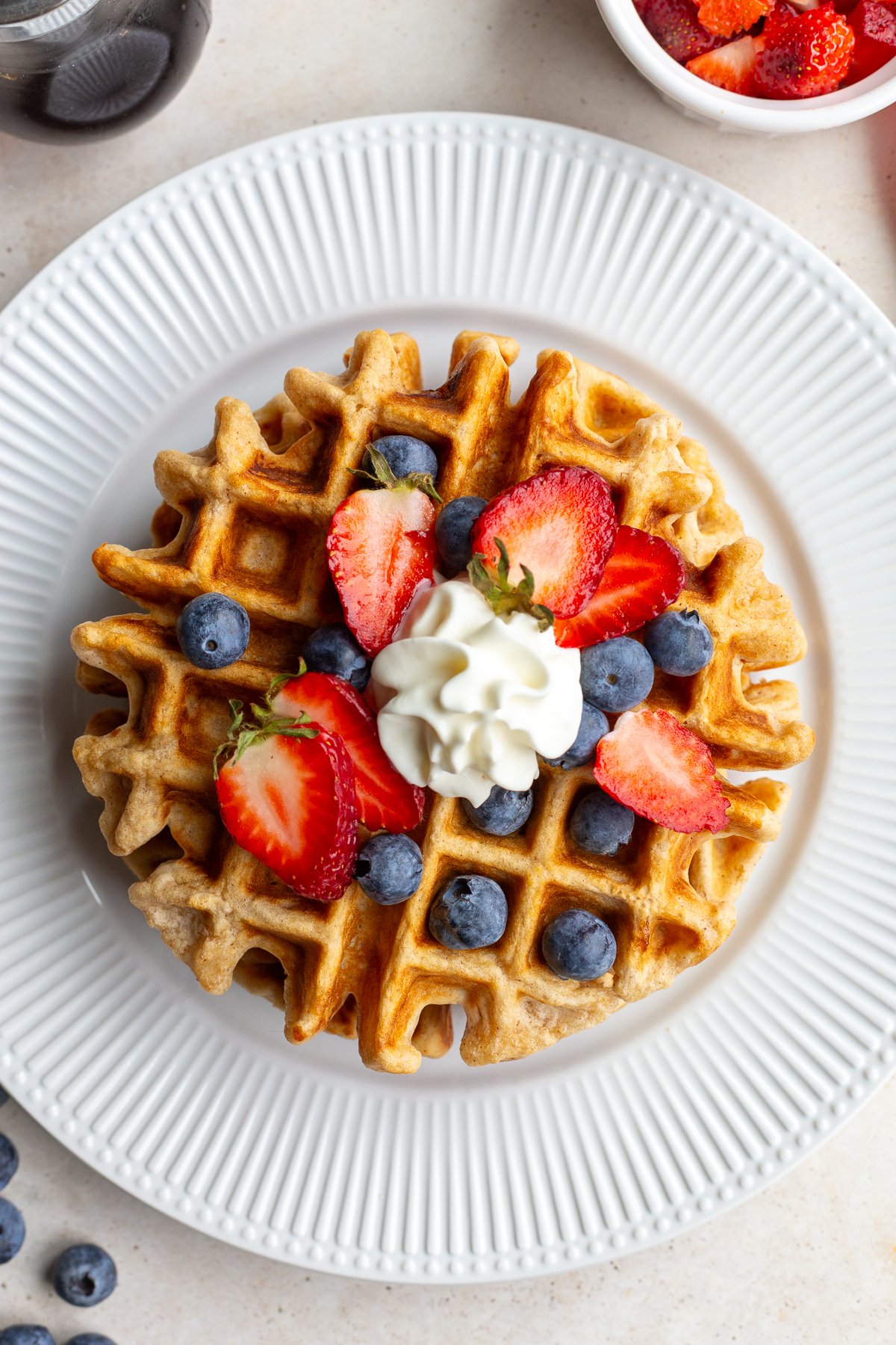 Waffle on a white plate topped with berries and whipped cream.