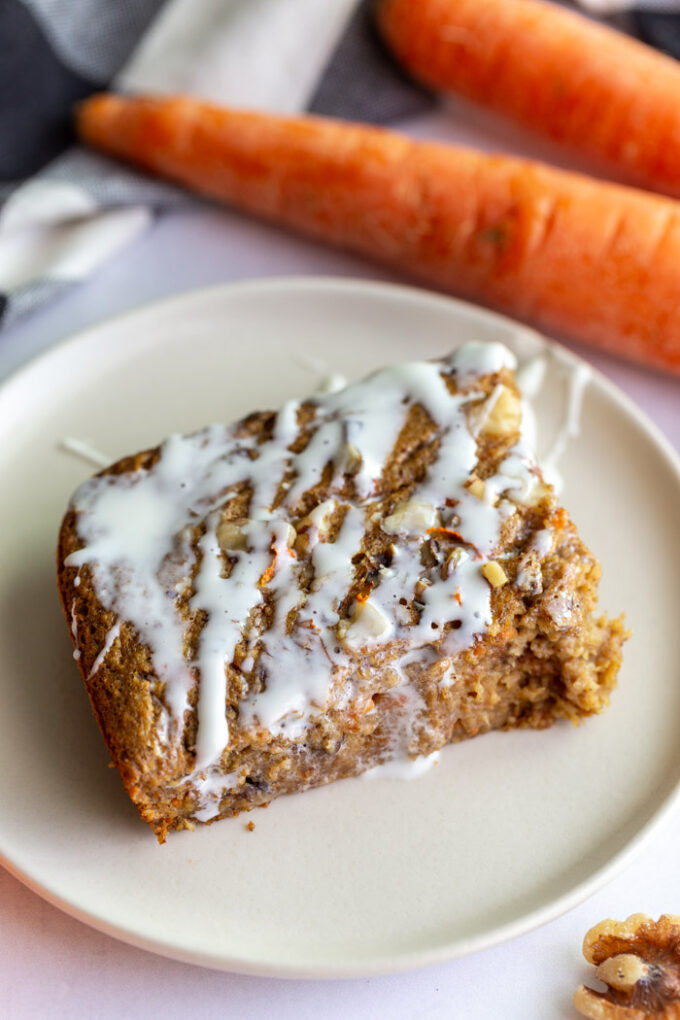 slice of carrot cake baked oats on a plate with cream cheese frosting drizzled on top. carrots in the background