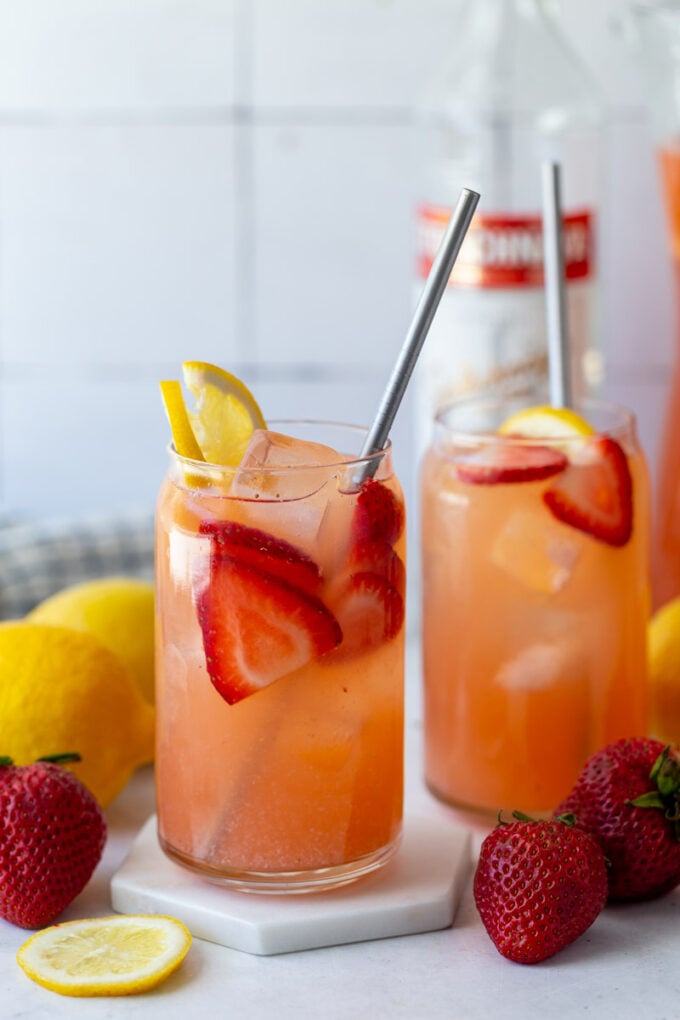 glass filled with strawberry lemonade vodka with a bottle of vodka in the background and garnished with fresh lemon and strawberries