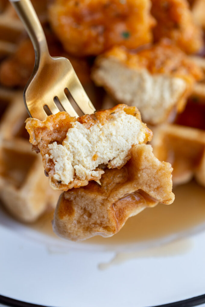 bite of vegan chicken and waffles on a fork with the rest of the dish behind in