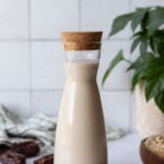 tall clear container of oat milk with fresh dates and oats around it and a fresh plant in the corner