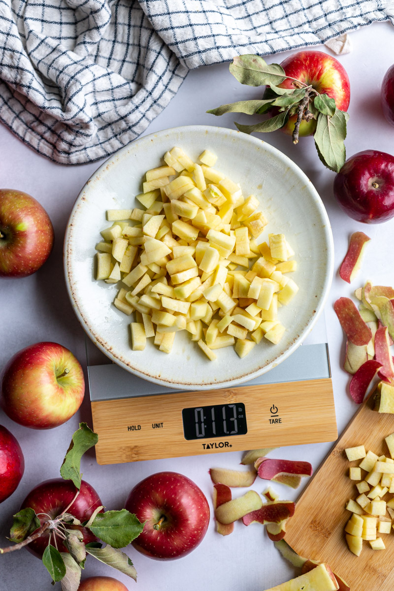 apple pieces on a plate on a kitchen scale with lots of apples and spple scrapes around the scene. towel in the corner and fresh apples around