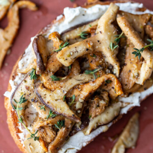 dark red plate with toast on top. Toast has goat cheese and sautéed mushrooms on top with fresh herbs