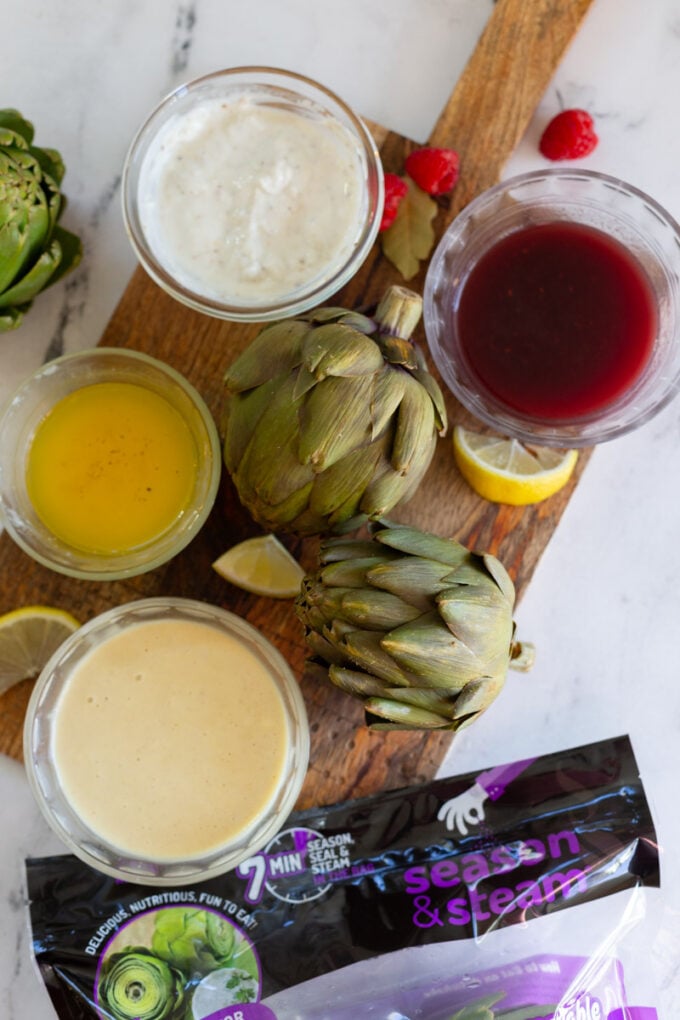 4 different artichoke dips on a cutting board with 2 steamed artichokes