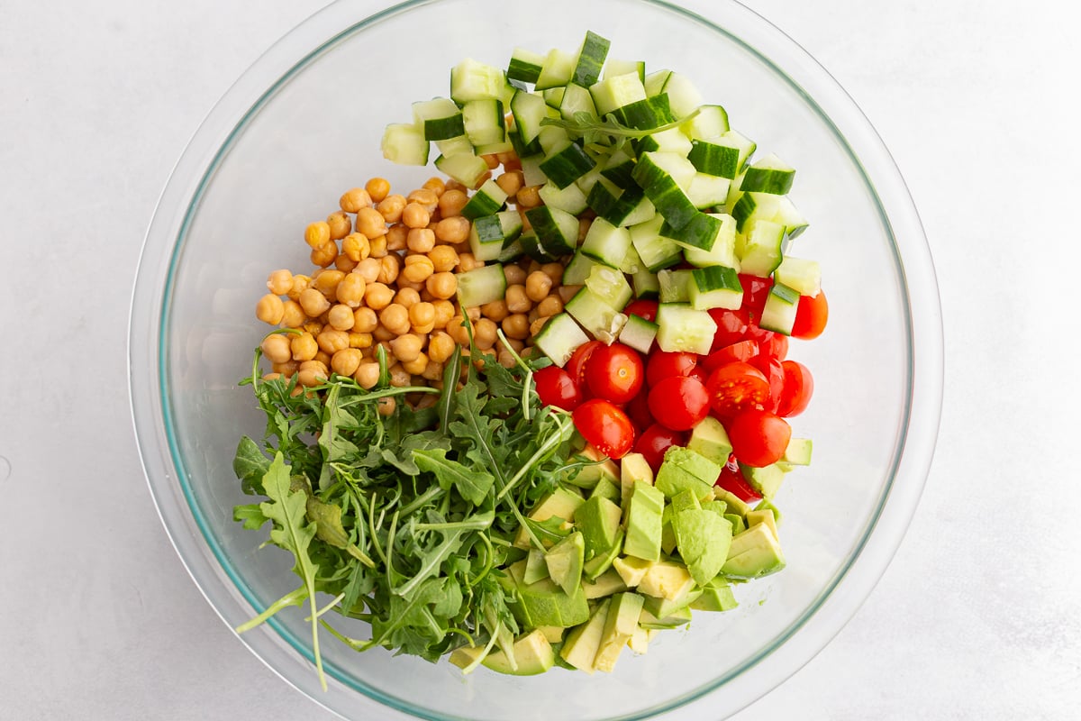 ingredients in a large bowl including chickpeas, arugula, tomatoes, diced cucumber, and avocado diced
