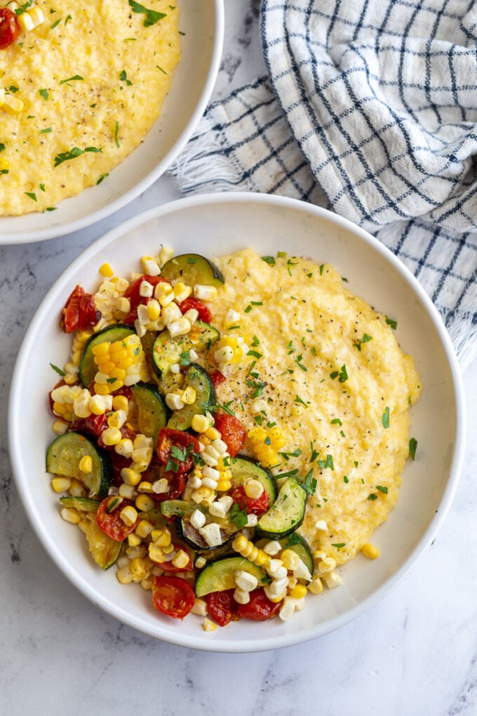 2 bowls of cheesy polenta topped with roasted zucchini, corn, and tomatoes and sprinkled with fresh herbs