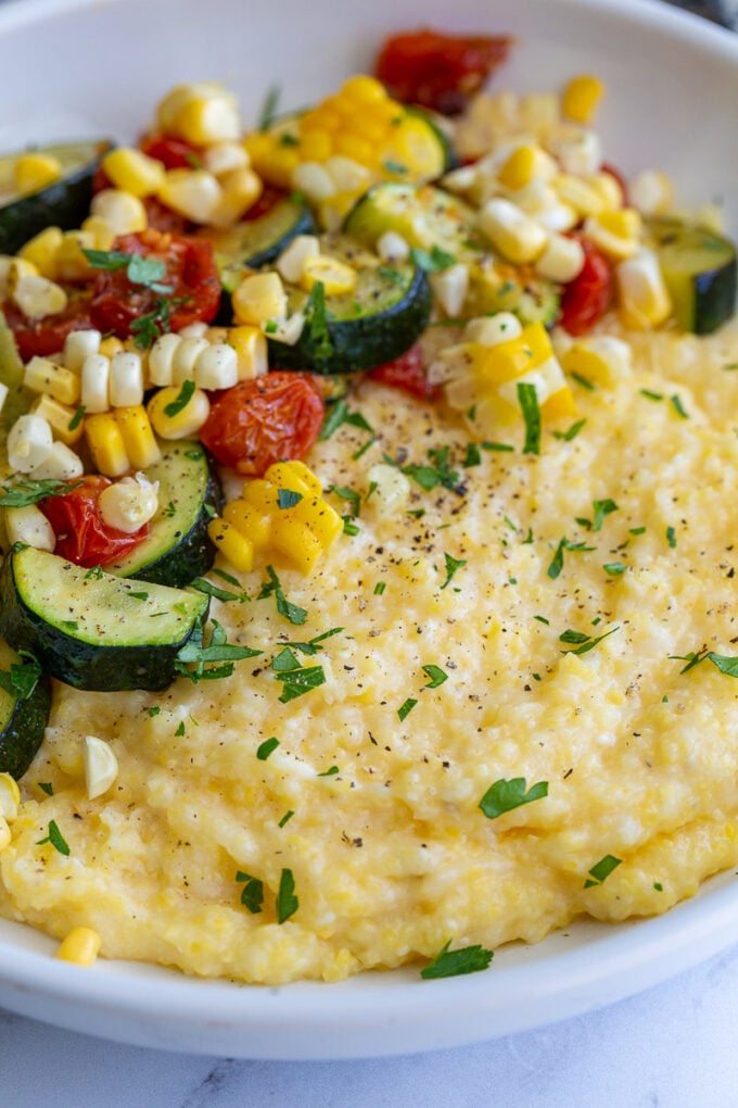 bowl of cheesy polenta topped with roasted zucchini, corn, and tomatoes and sprinkled with fresh herbs