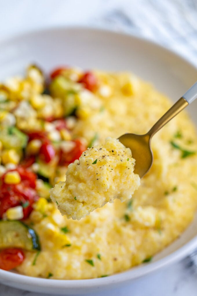 bowl of cheesy polenta topped with roasted zucchini, corn, and tomatoes and sprinkled with fresh herbs with a fork holding up a bite of the polenta