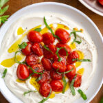 Whipped ricotta cheese in a big bowl that's topped with bright red burst cherry tomatoes and green basil ribbons and a drizzle of olive oil