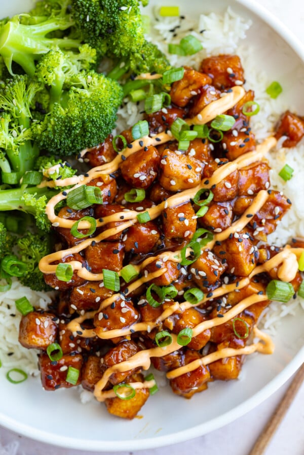 honey garlic tofu in a bowl on top of white rice with cooked broccoli next to it. It's covered in green onion, sesame seeds, and a drizzle of spicy mayo sauce.
