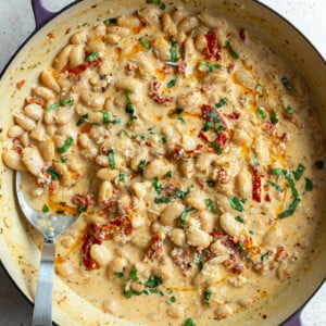 large skillet with creamy butter beans and sundried tomtoes in it known as "marry me butter beans". topped with fresh basil., there's a large spoon on the side