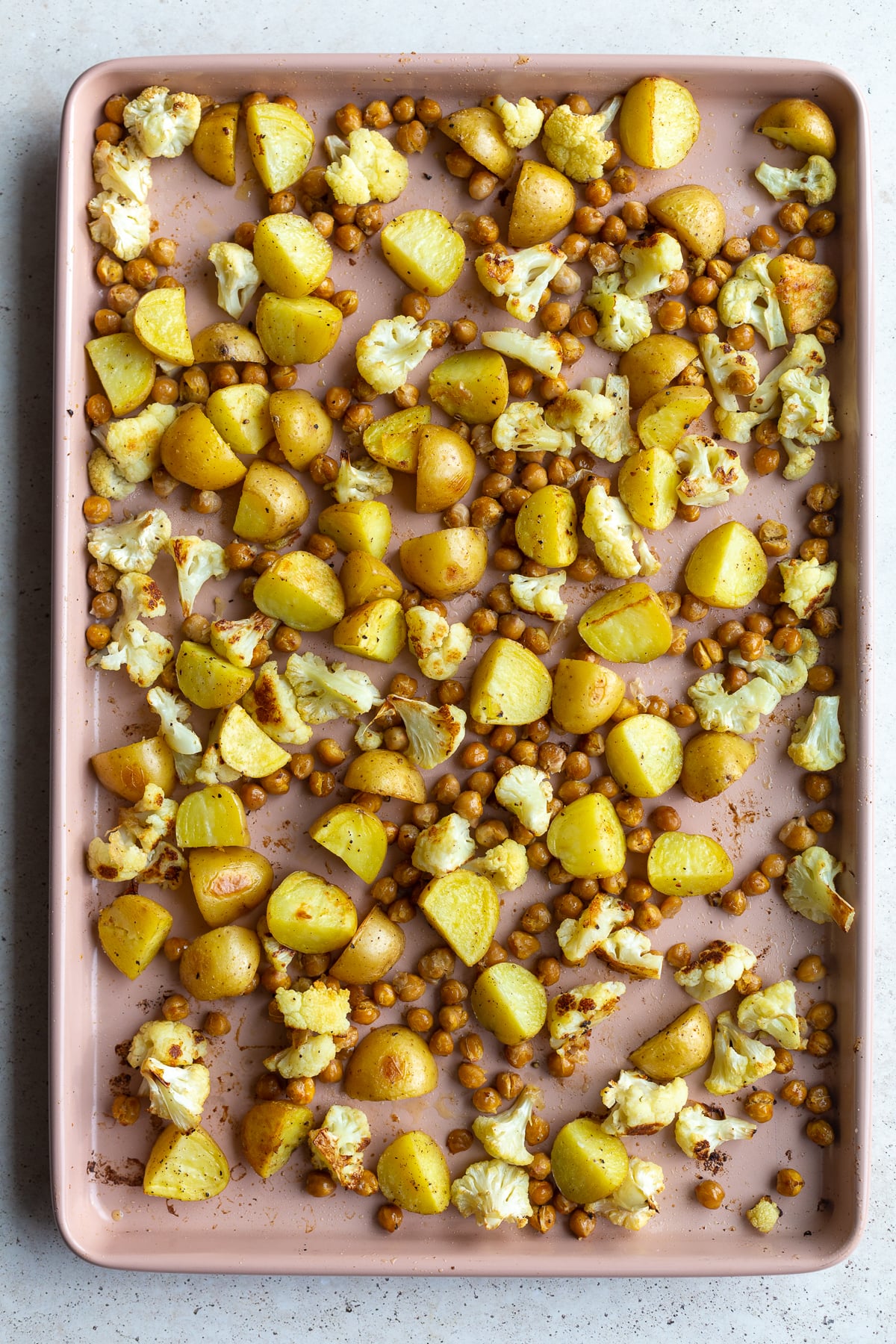 A light pink baking sheet laden with uniformly diced potatoes, chickpeas, and cauliflower florets, ready for roasting.