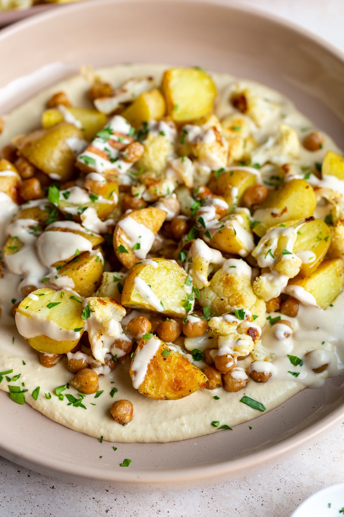A hearty and colorful veggie bowl featuring roasted potatoes, chickpeas, and cauliflower on a generous bed of creamy hummus, topped with a luscious tahini sauce drizzle.
