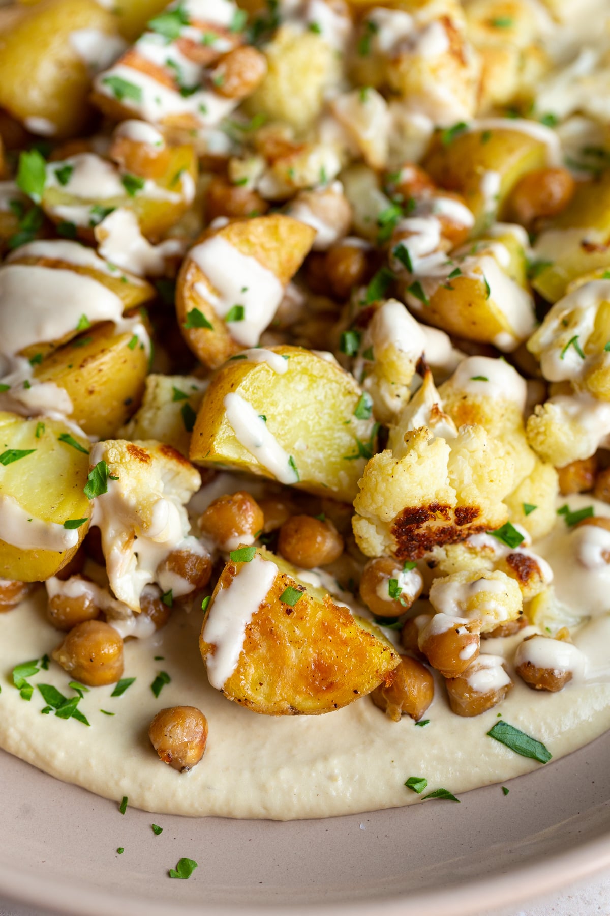 Close-up of a veggie bowl showcasing roasted potato, chickpeas, and cauliflower on a bed of creamy hummus, elegantly drizzled with a light tahini sauce.