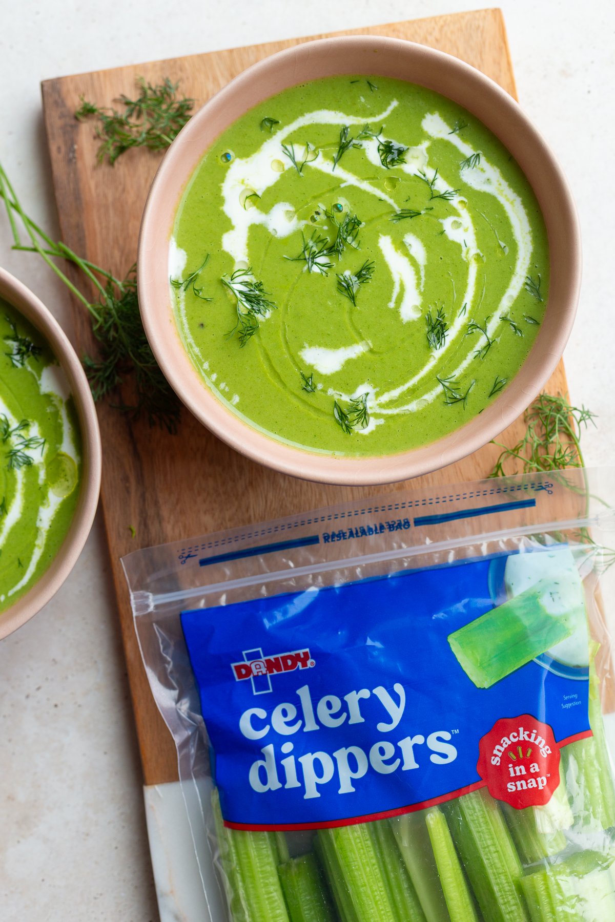 green goddess soup that's vibrant green in a pink bowl with a cream drizzle. there's a bag of "celery dippers" in the corner of the shot