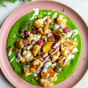 green goddess veggie bowls with a creamy green sauce smeared on a pink plate + roasted cauliflower, radish, baby potatoes, and chickpeas on top. The whole plate is drizzled with a white tahini sauce