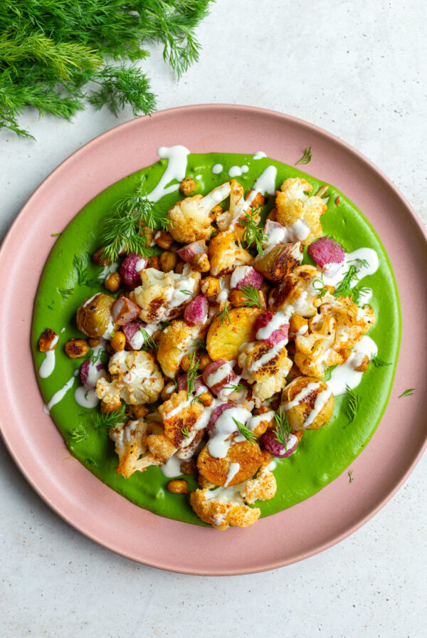 green goddess veggie bowls with a creamy green sauce smeared on a pink plate + roasted cauliflower, radish, baby potatoes, and chickpeas on top. The whole plate is drizzled with a white tahini sauce