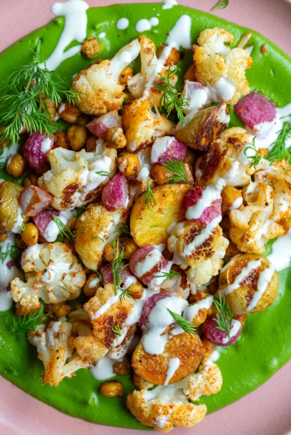 green goddess veggie bowls with a creamy green sauce smeared on a pink plate + roasted cauliflower, radish, baby potatoes, and chickpeas on top. The whole plate is drizzled with a white tahini sauce