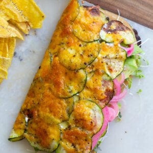 cheesy zucchini wrap stuffed with vegan meat, red onion, avocado and tomato. The wrap is on a plate with chips