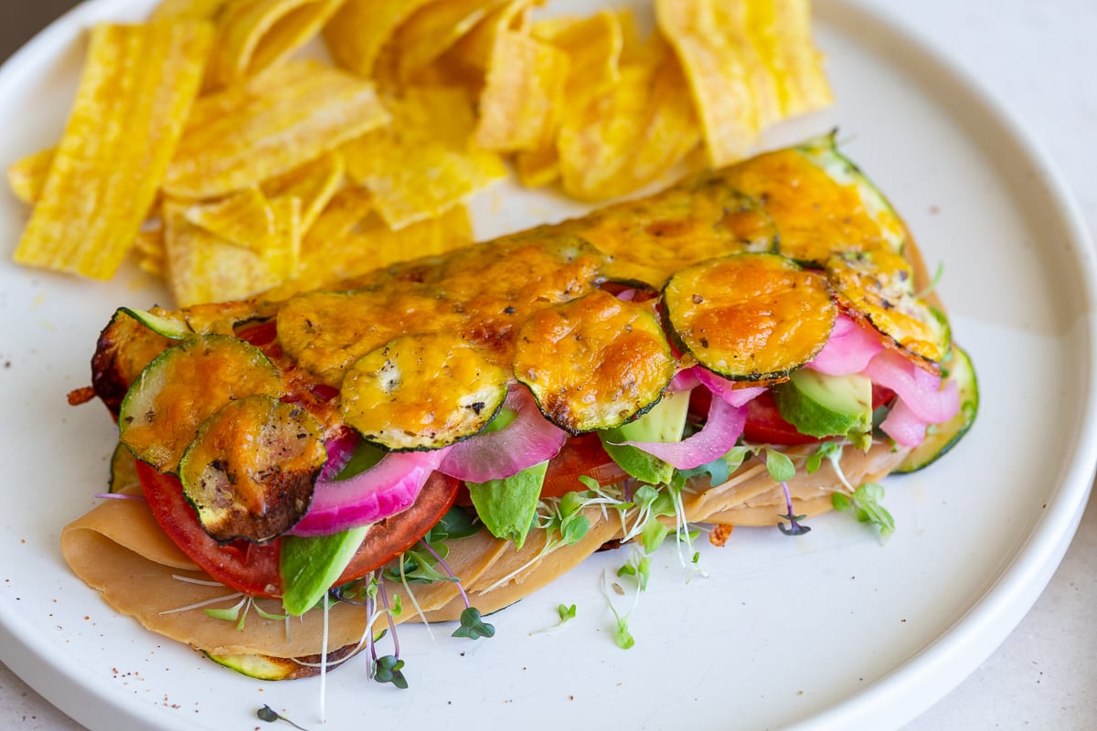 cheesy zucchini wrap stuffed with vegan meat, red onion, avocado and tomato. The wrap is on a plate with chips