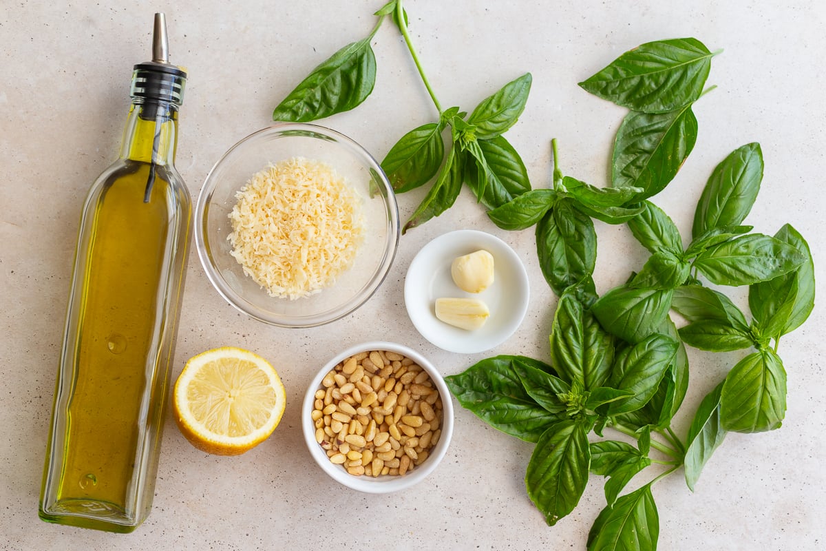 ingredients laid out to make homemade pesto including olive oil, parmesan, toasted pinenuts, fresh basil, lemon, and garlic