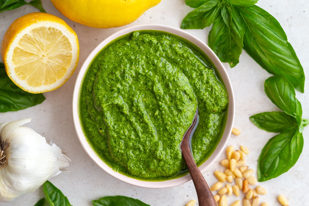 homemade pesto recipe - a bowl of pesto with a wooden spoon in it and lemons and fresh basil in the corners of the shot