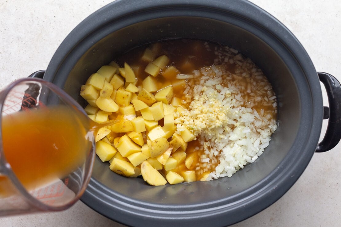 potatoes, onion and garlic in a crockpot with broth being poured in to make slow cooke potato soup