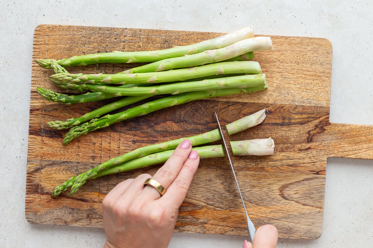 asparagus on a wood cutting board with a hand cutting off the woody ends of the asparagus