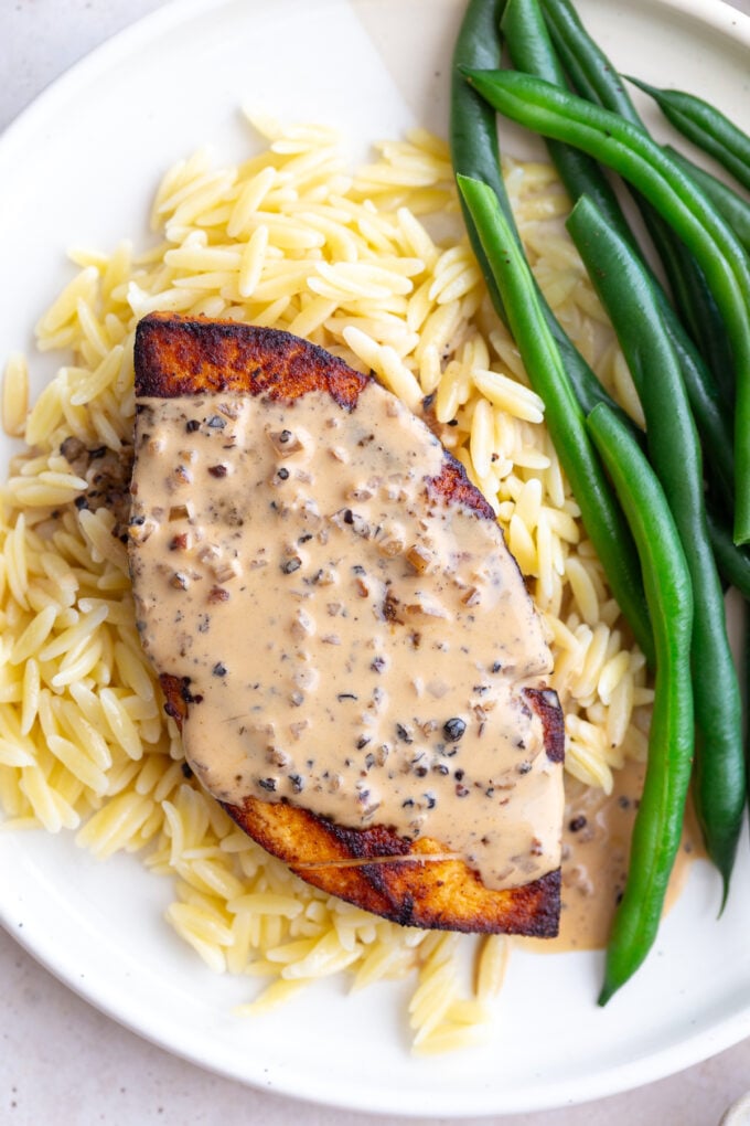 tofu "steak" laying on a bed of orzo, next to green beans, and covered in a creamy peppercorn sauce