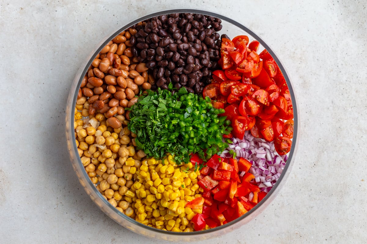 bowl filled ingredients that are neatly sectioned out and not mixed together yet including: pinto beans, black beans, chickpeas, tomato, onion, jalapeno, coen and bell pepper