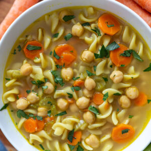 chickpea noodle soup in a large white bowl with fresh carrots and celery around the shot