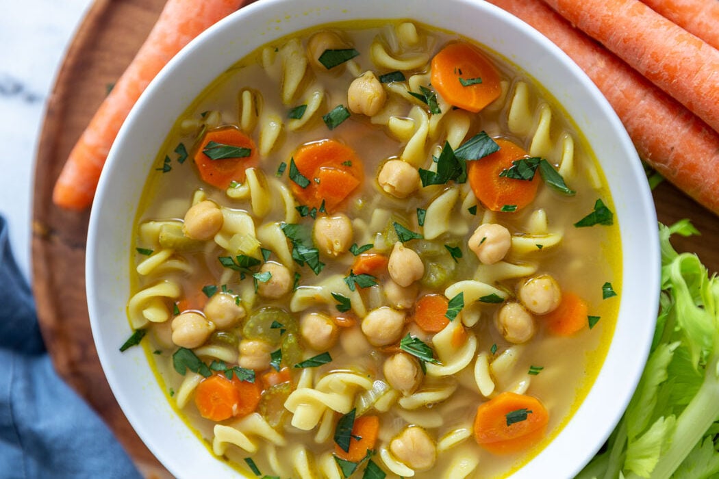 chickpea noodle soup in a large white bowl with fresh carrots and celery around the shot