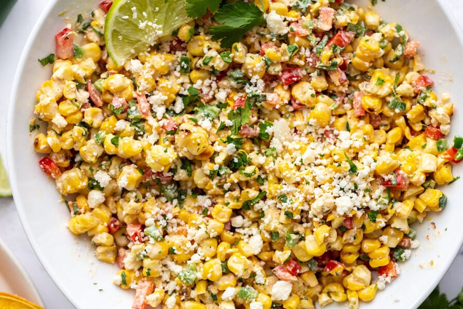 large white bowl of mexican street corn salad topped with cotija cheese and a lime with cilantro in the scene
