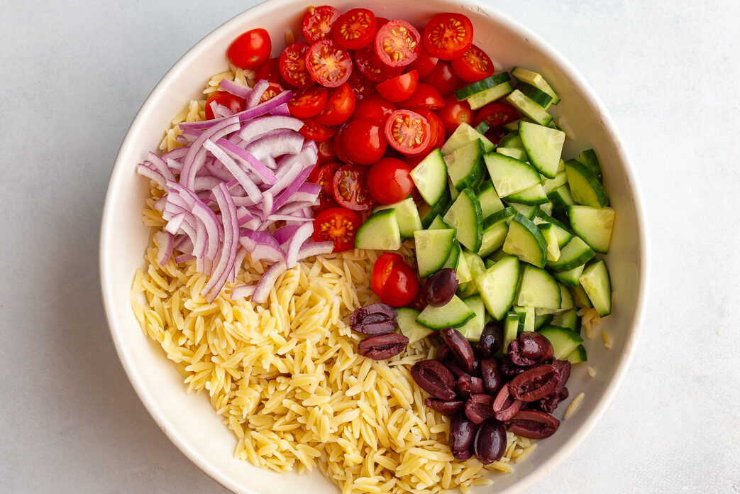 orzo salad in a bowl before being mixed together including cooked orzo, cherry tomatoes, cut cucumber, sliced red onion, and sliced olives