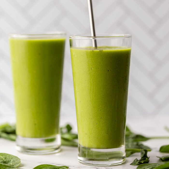2 glasses filled with green smoothie on a marble board with spinach sprinkled al around