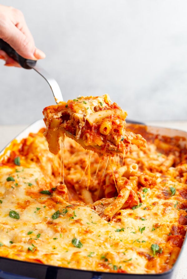 baked ziti in a casserole dish with a hand using a scoop to pull some of the baked ziti out of the pan