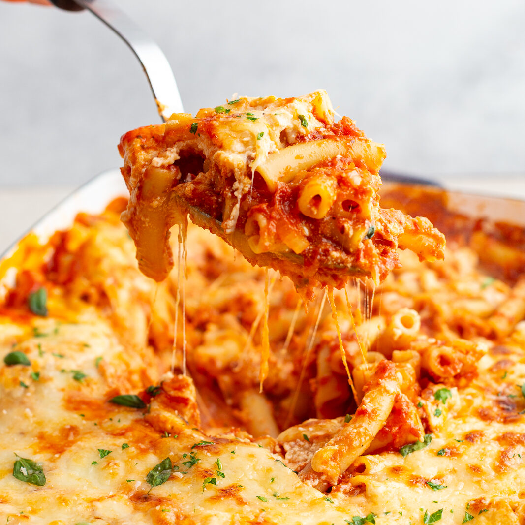 baked ziti in a casserole dish with a hand using a scoop to pull some of the baked ziti out of the pan