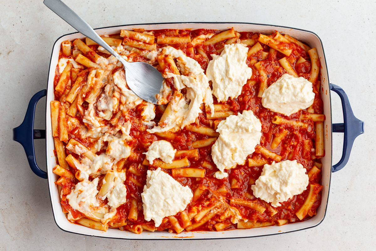 baked ziti being assembled where you see the marinara covered ziti in the casserole dish topped with swirled ricotta