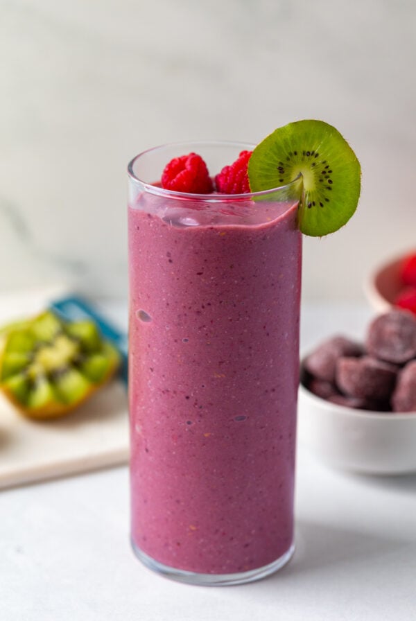 acai smoothie in a tall glass topped with raspberry and kiwi with more kiwi in the background