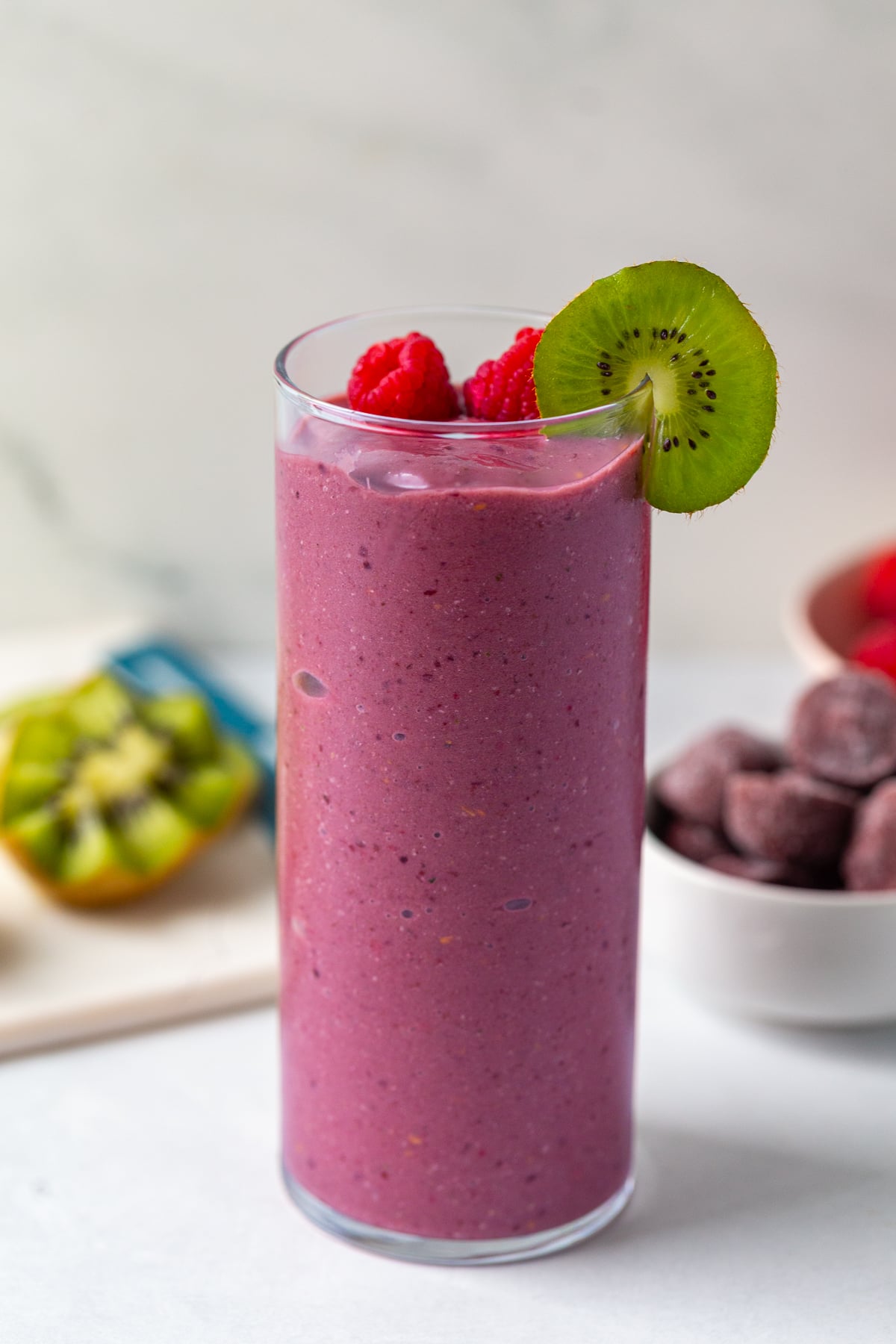 acai smoothie in a tall glass topped with raspberry and kiwi with more kiwi in the background