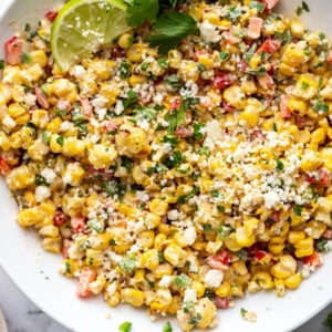 large white bowl of mexican street corn salad topped with cotija cheese and a lime with cilantro in the scene