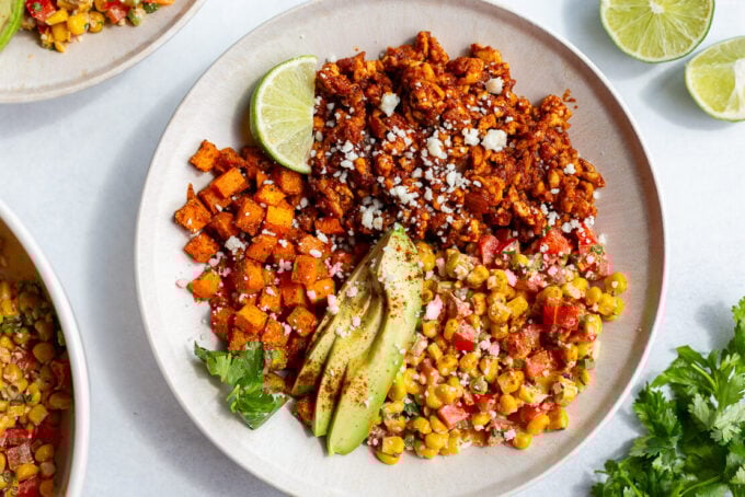 street corn tofu bowls with roasted sweet potato, ground tofu "meat", sliced avocado, lime wedge, and street corn salad in a bowl