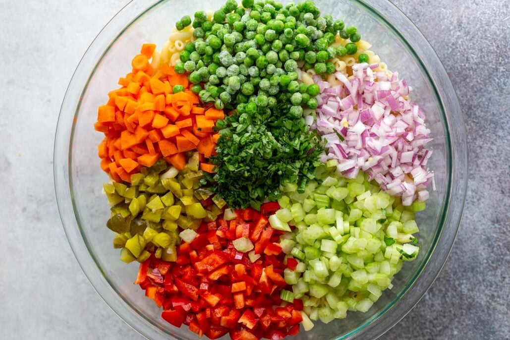 ingredients in a large bowl to make easy macaroni salad including frozen peas, chopped carrots, chopped red onion, chopped red bell pepper, chopped pickles, and chopped celery and parsley