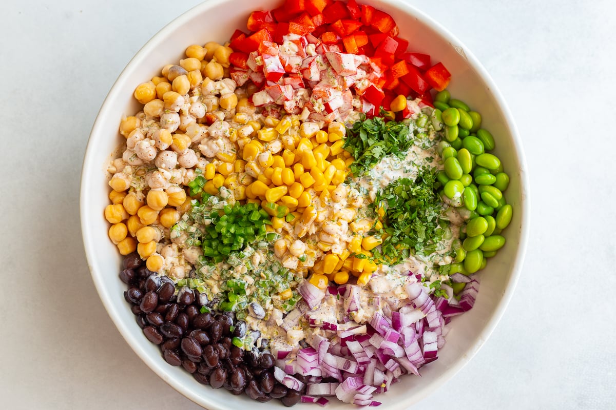 street corn salad veggies in a large bowl for dense beans salad including corn, beans, cilantro, jalapeno, red onion, and edamame. Topped with a cream dressing drizzled over the top