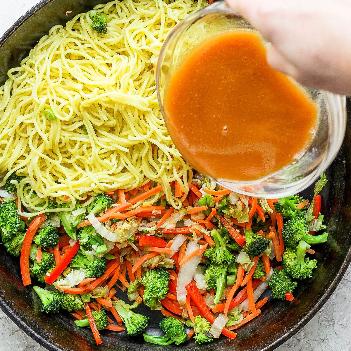 veggies and noodles in a cast iron skillet with sauce being poured in