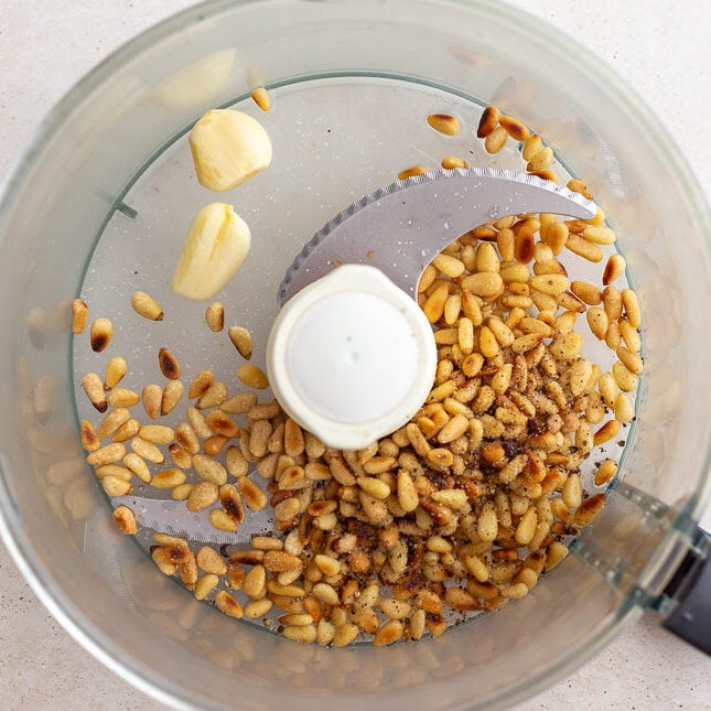 how to make pesto step 2: picture of a food processor with toasted pinenuts, lemon juice, and garlic cloves in it