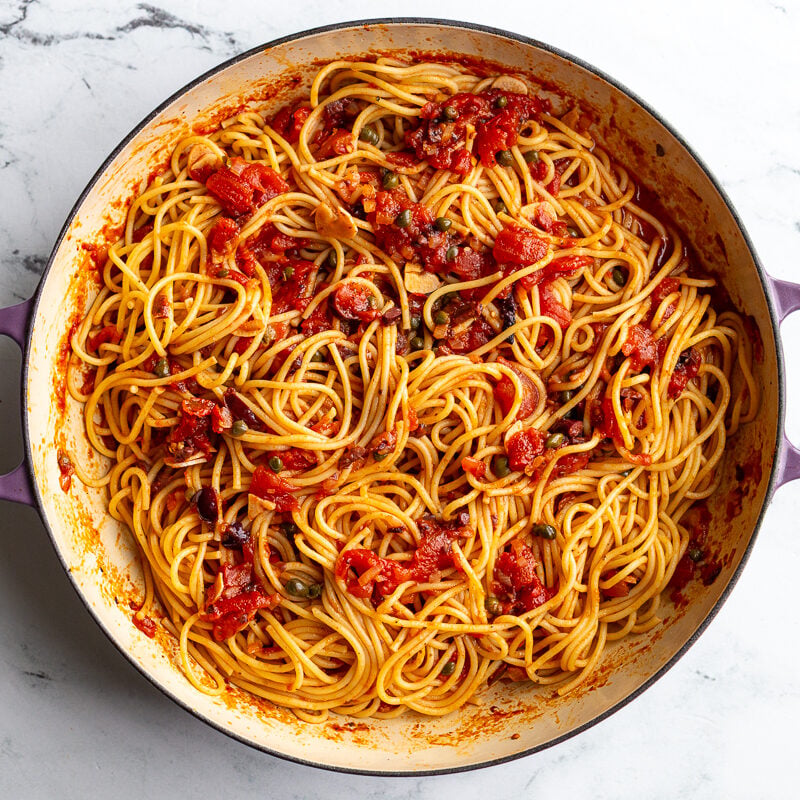 Spaghetti puttanesca tossed together in a skillet with sauce, olives, and capers.