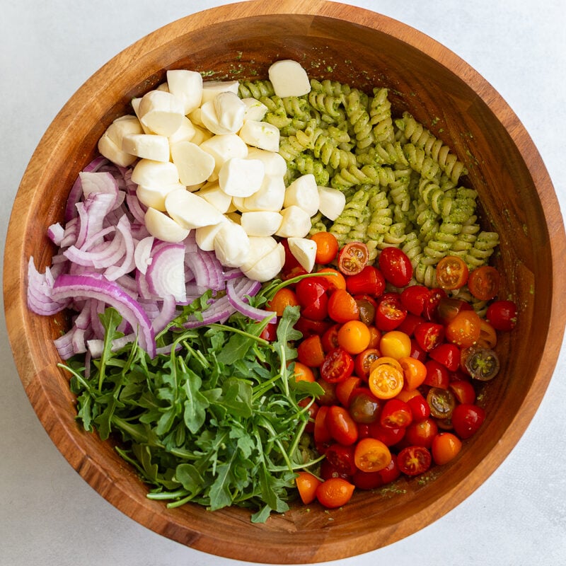 pesto pasta in a large wood bowl with mozzarella, tomatoes and arugula on top before being mixed in