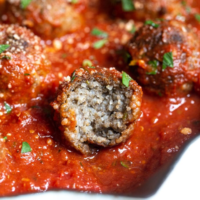 up close shot of vegetarian meatballs in a skillet with tomato sauce and sprinkled with vegan cheese and parsley. one with a bite taken out