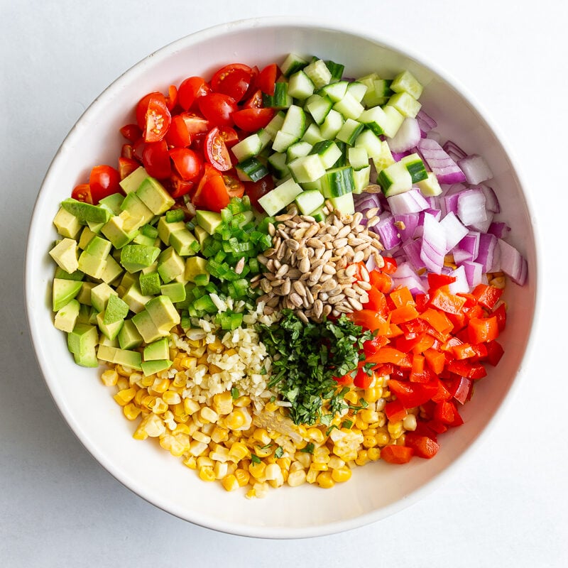 grilled corn salad ingredients in a large white bowl including corn, sunflower seeds, bell pepper, cucumber, red onion, avocado etc
